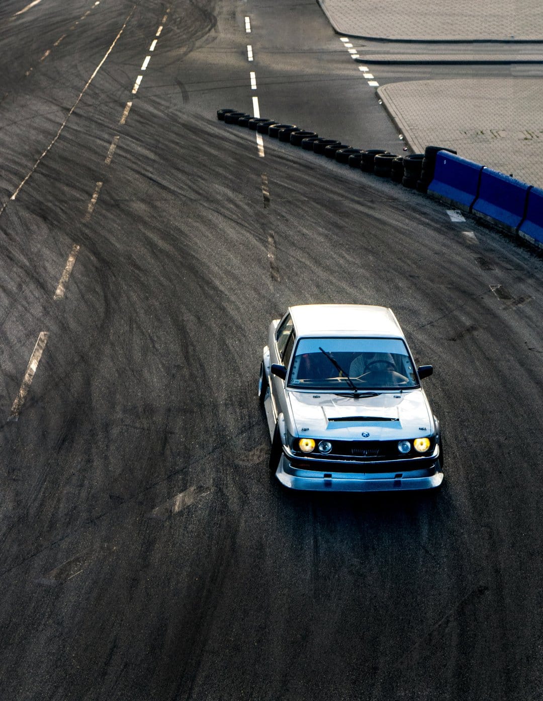 Silver car drifting on a dark asphalt track with rubber marks, blue barriers, and white dashed boundary lines.