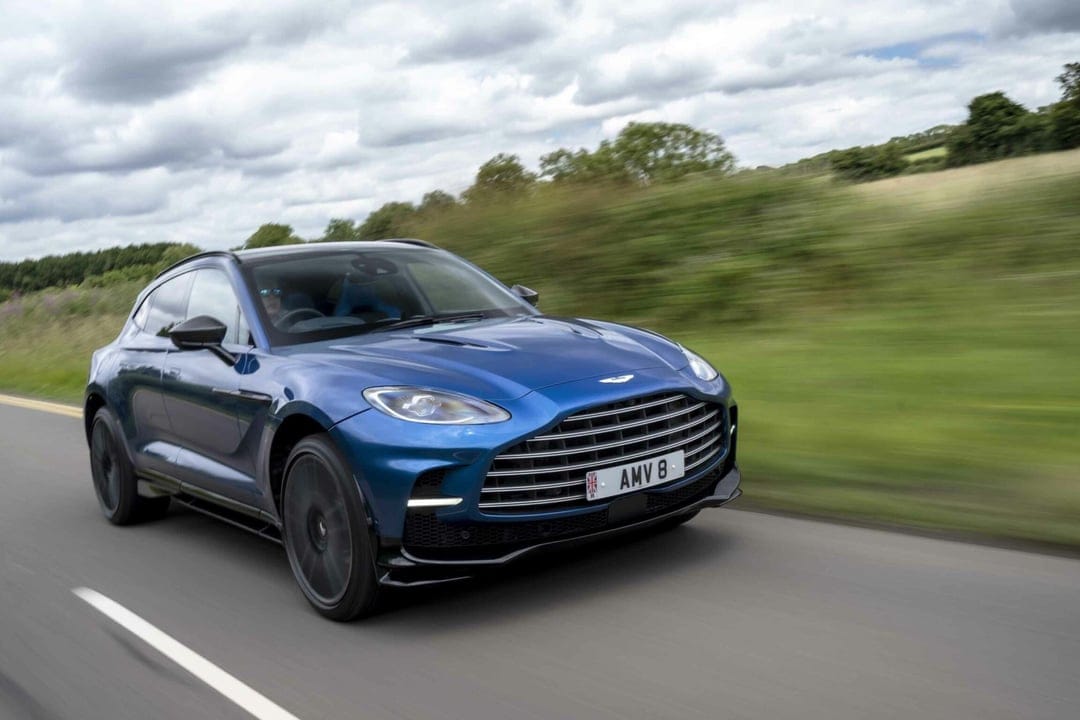 Blue Aston Martin DBX luxury SUV driving on a paved road with motion blur and a cloudy sky background.