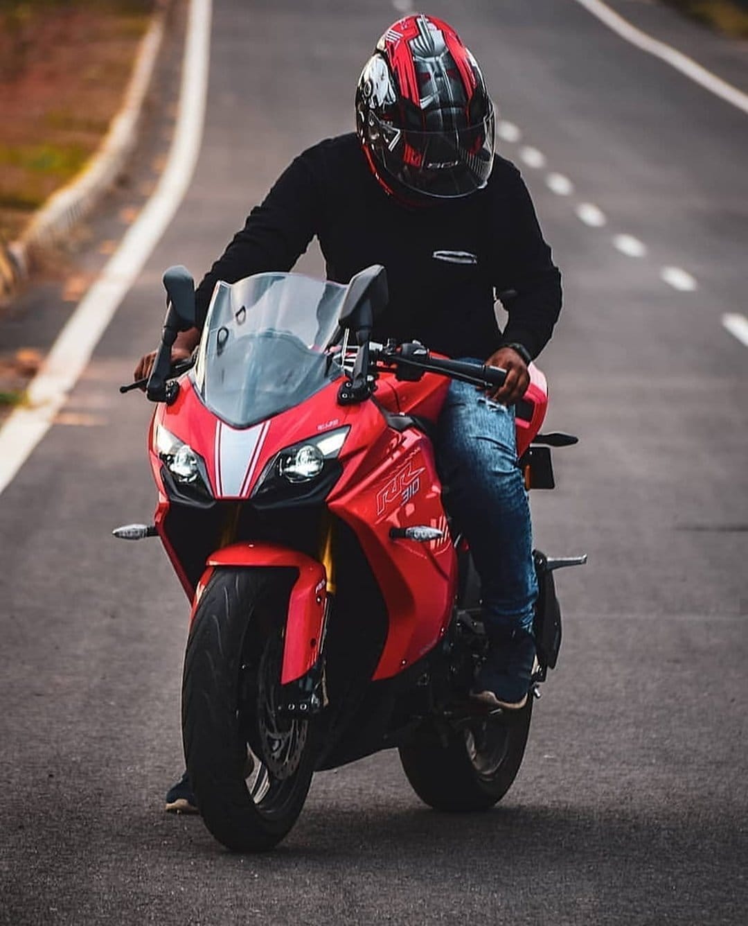 Red sportbike rider wearing a black shirt and patterned helmet travels along a paved highway with greenery.
