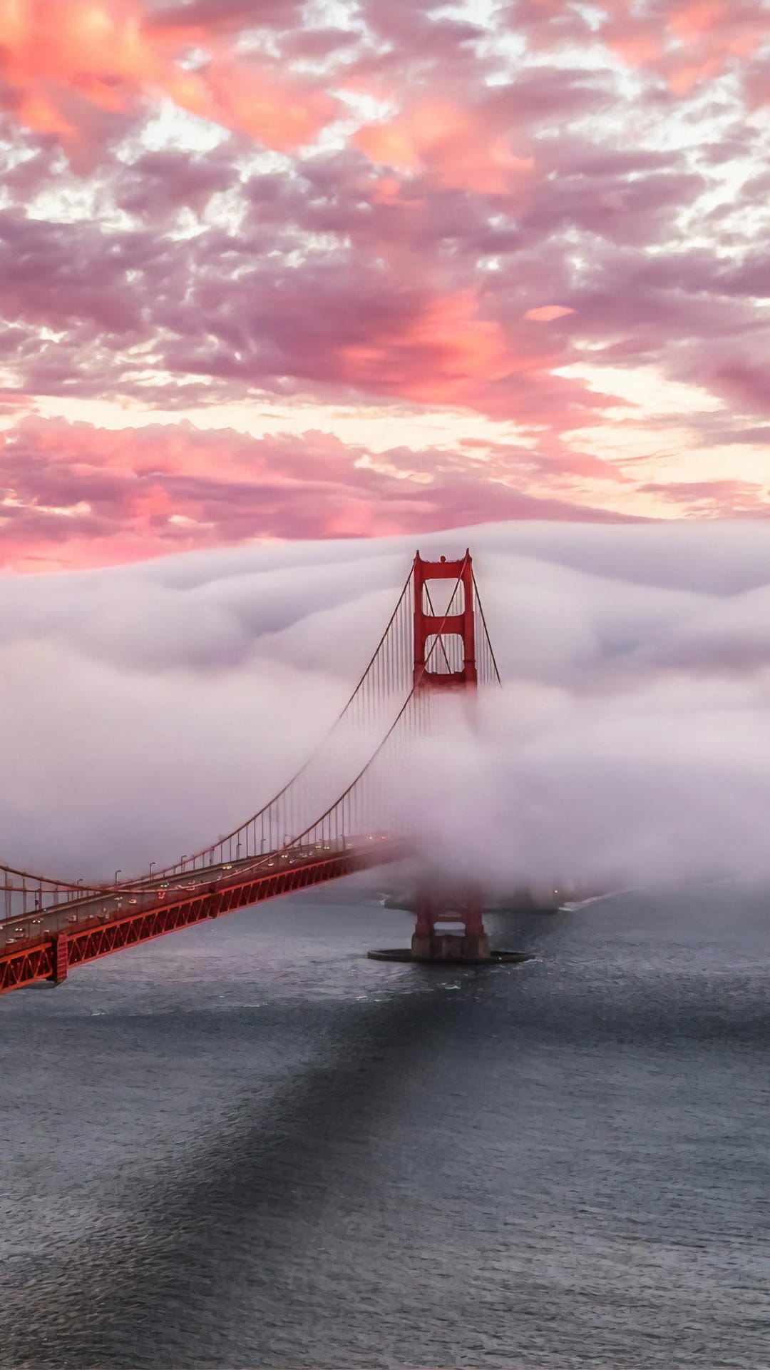 Red Golden Gate Bridge towers pierce through dense pink mist over dark water during a purple sunset.