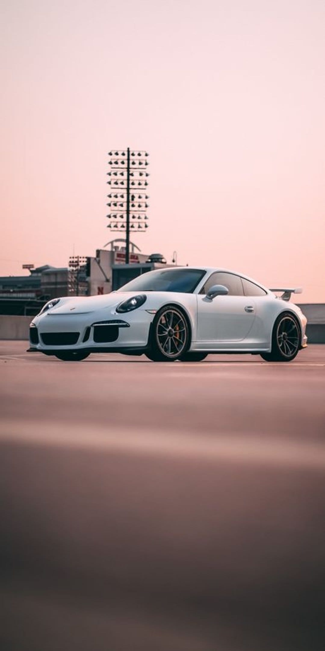White Porsche 911 GT3 sports car on textured asphalt under tall stadium lights during a soft orange sunset.