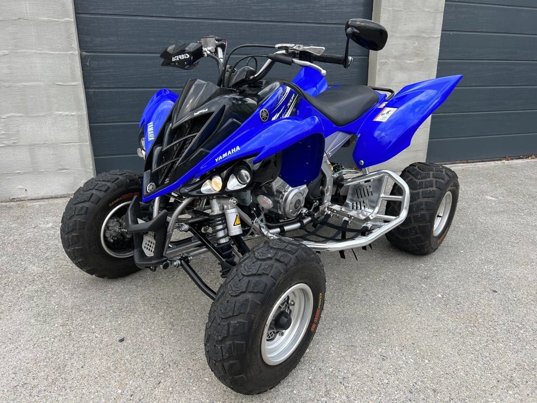 Blue Yamaha Raptor 700R sport ATV with knobby tires parked on a paved driveway against grey garage doors.