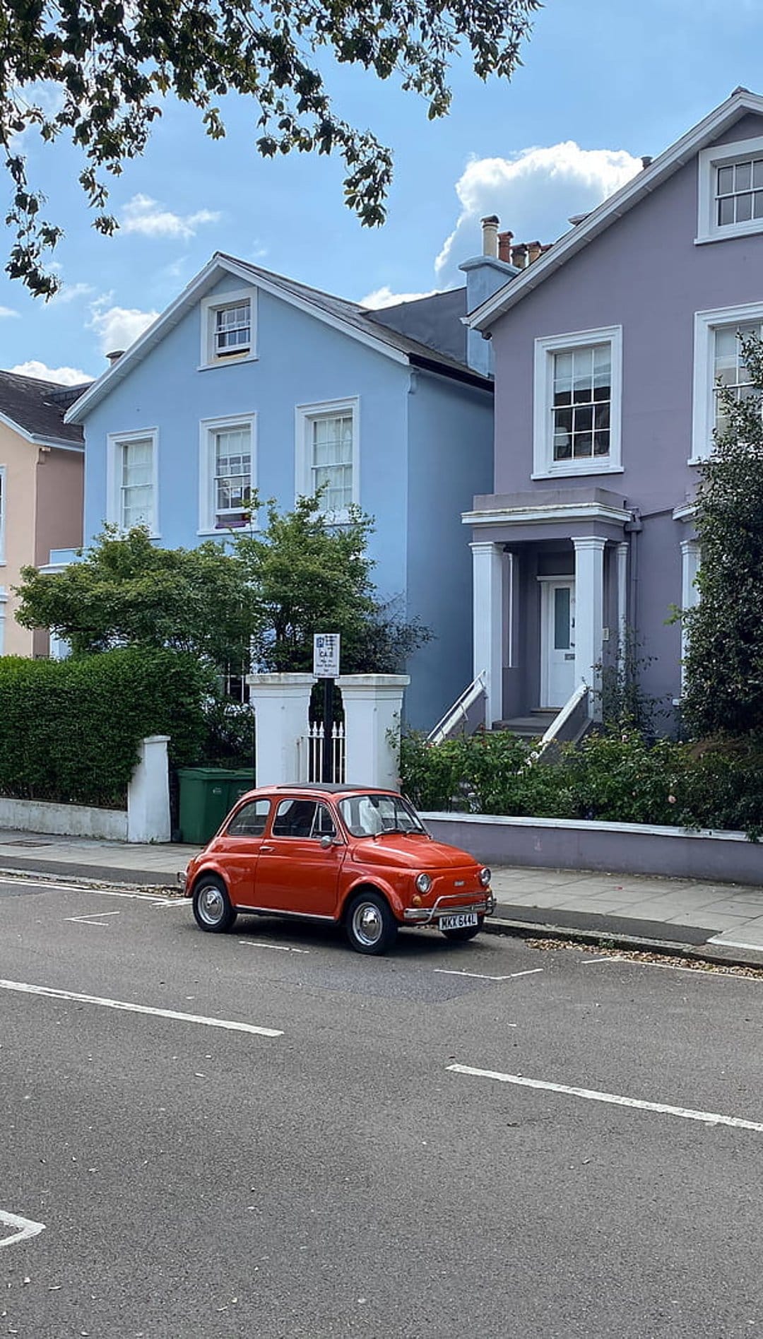 Orange Fiat 500 parked on a street with powder blue and lilac houses under a bright sky with white clouds.