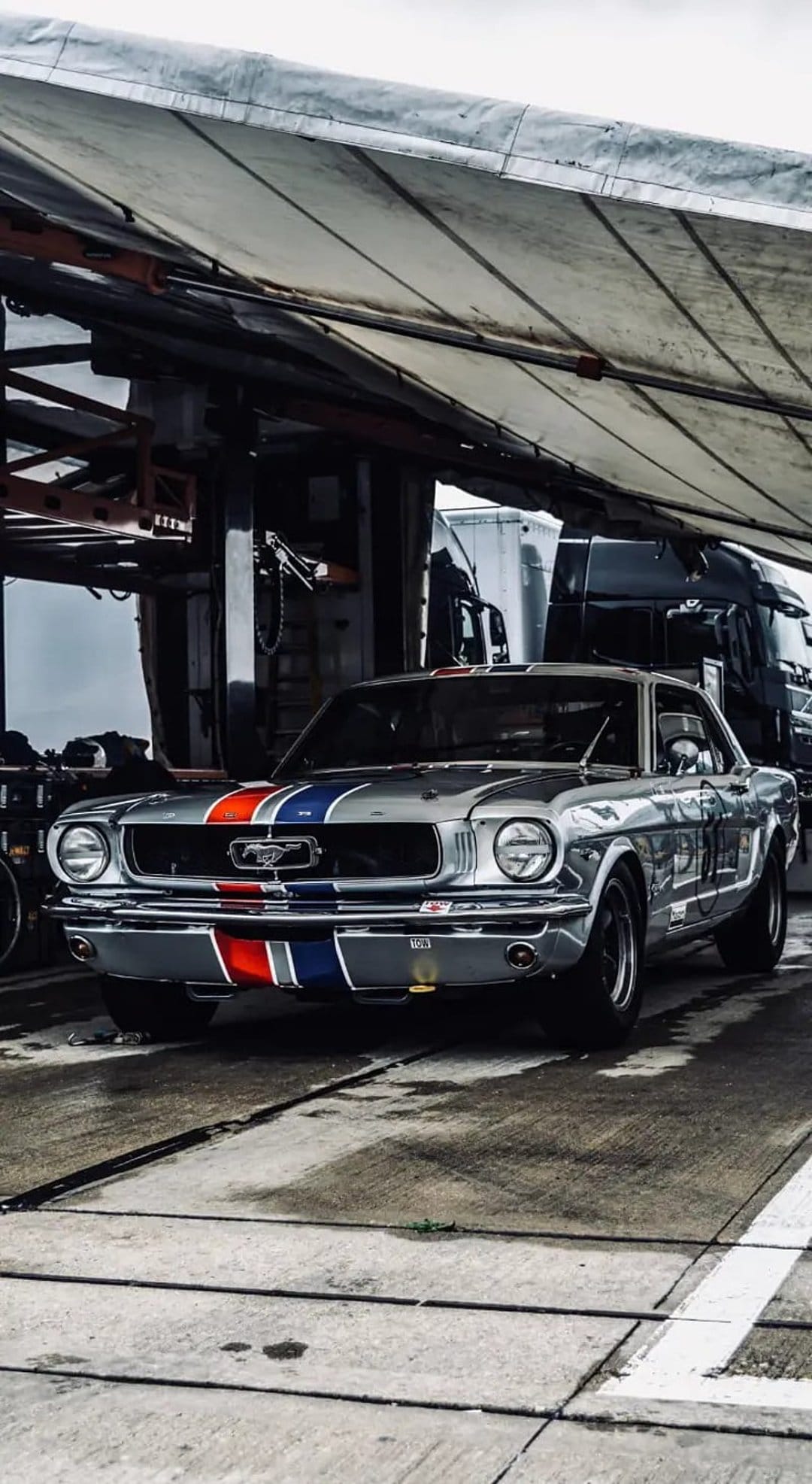 Silver Ford Mustang GT with red and blue racing stripes parked on wet asphalt under an industrial canopy.