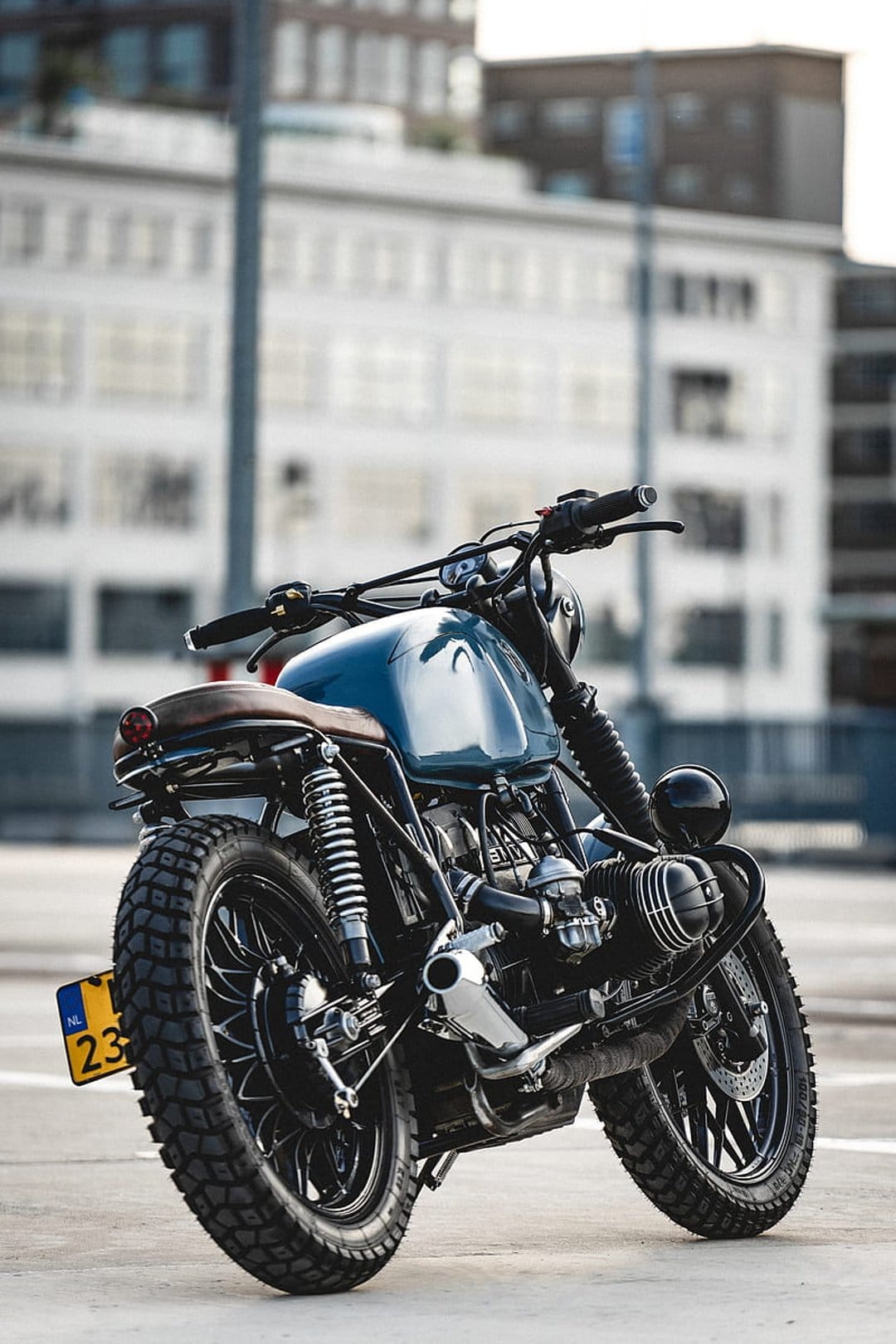 Blue BMW motorcycle with a brown leather seat and knobby tires parked in front of a blurred modern glass building.