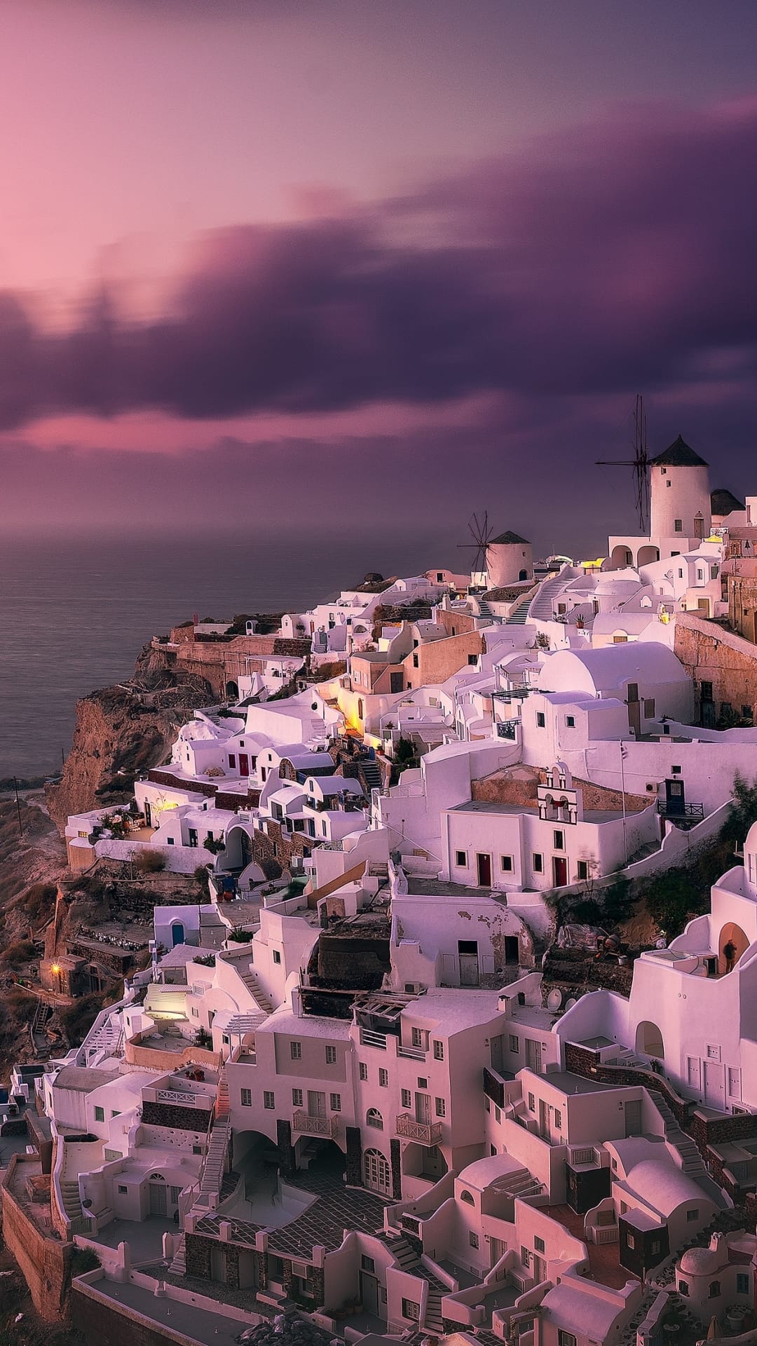 White-washed Santorini buildings and windmills sit atop steep cliffs under a pink and purple sunset sky.