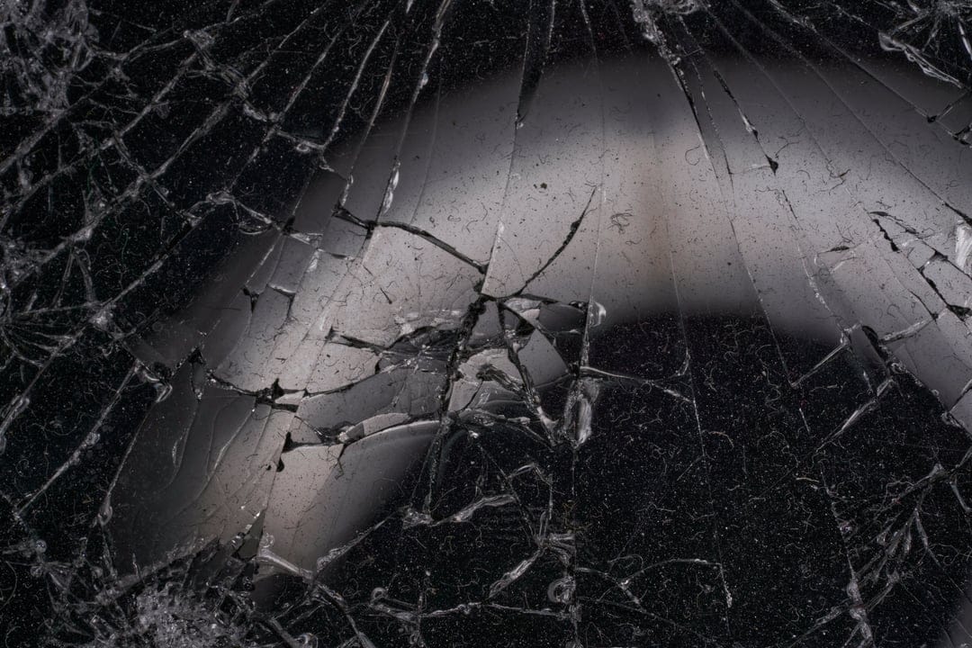 Intricate web of cracks on a dark shattered glass surface with sharp reflective edges and deep shadows.