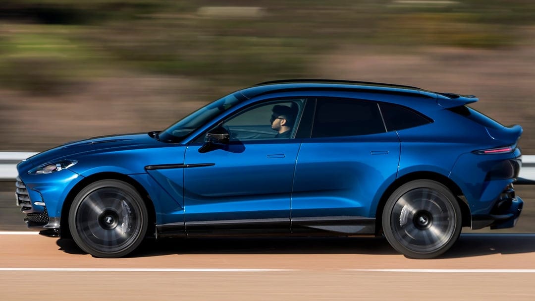 Side view of a blue Aston Martin DBX707 driving fast with motion blur background and spinning wheels.