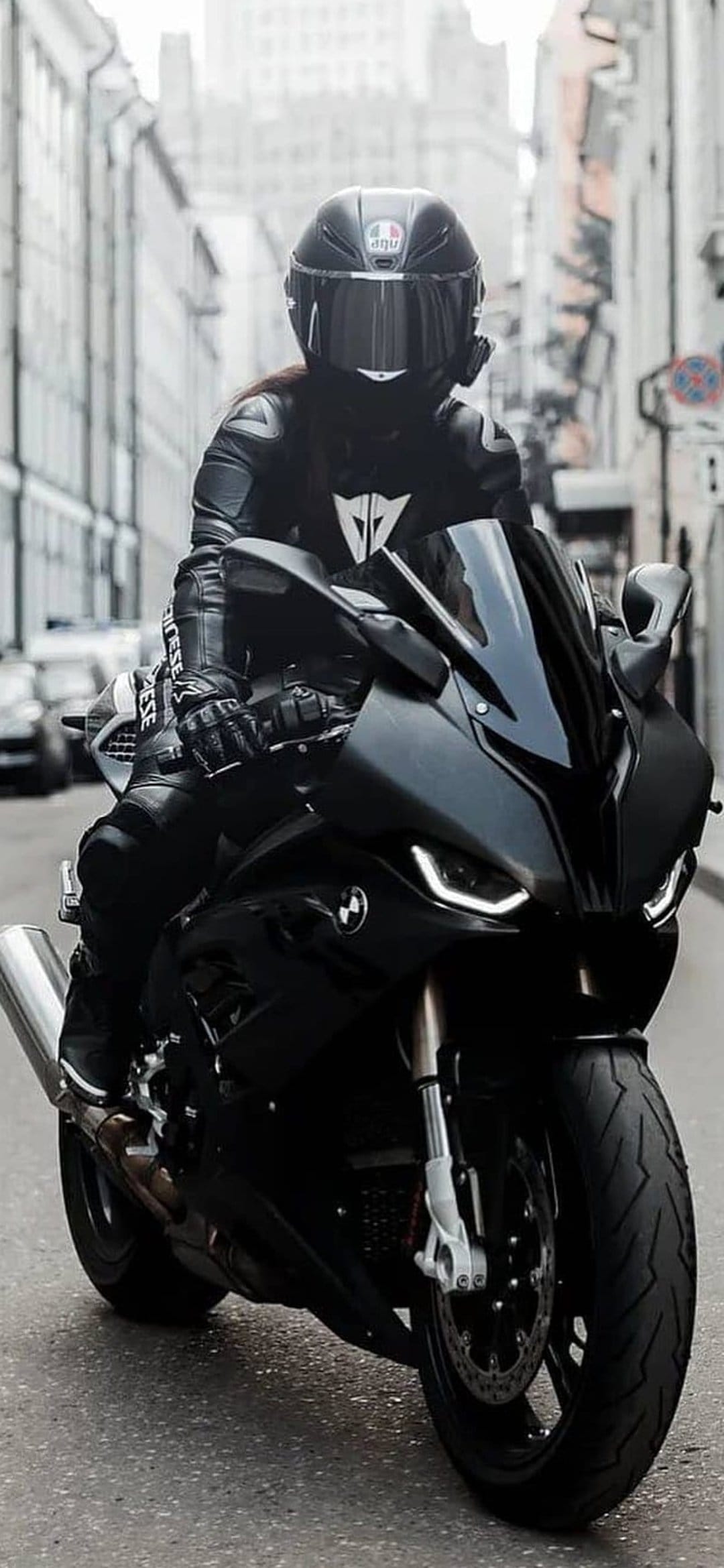 Motorcyclist in black leather gear sits on a sleek sportbike with glowing LED headlights on an urban city street.