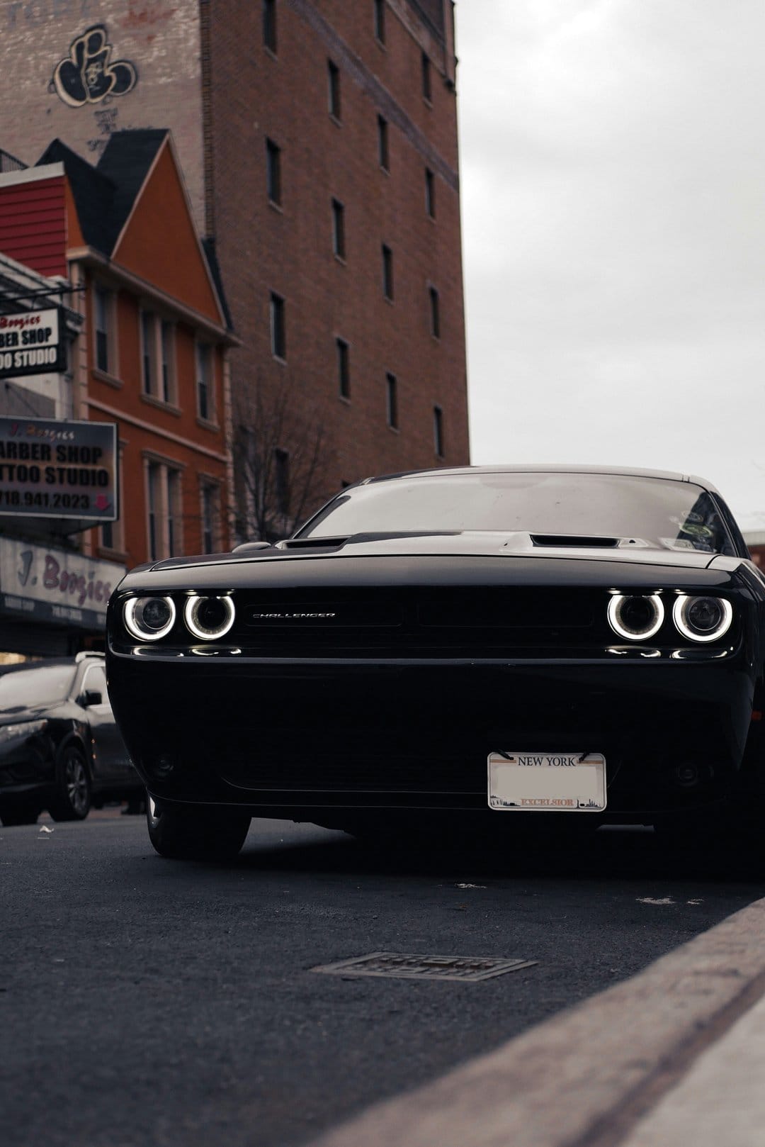Black Dodge Challenger muscle car with glowing halo headlights parked on a city street beside a brick building.