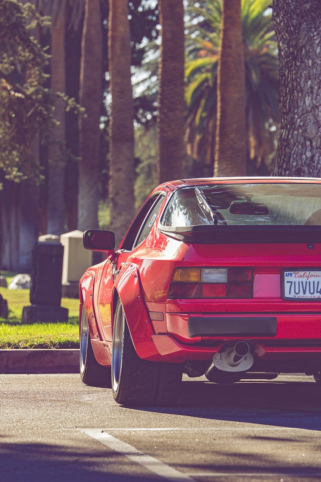 Red widebody Porsche 944 rear view with a large exhaust pipe and tall palm trees in a soft outdoor setting.