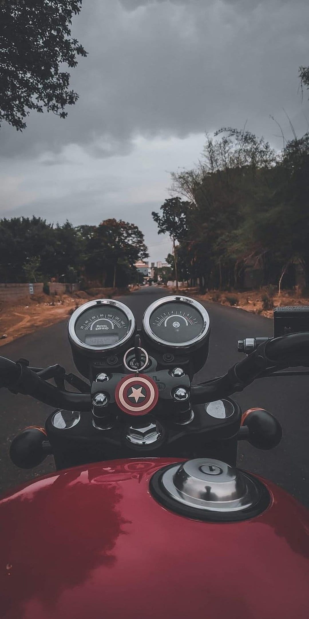Motorcycle dashboard with analog meters and a Captain America keychain on a misty road trip background.