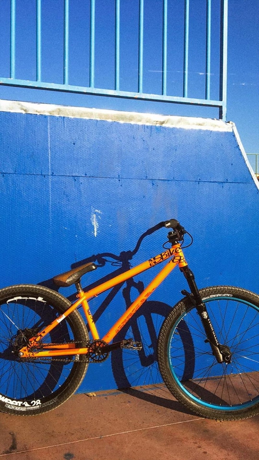Orange dirt jump bike with black handlebars leaning on a blue concrete wall under bright sunlight at a skate park.