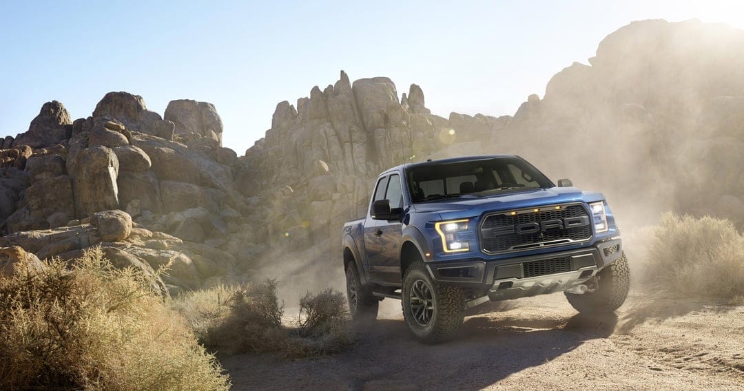 Blue Ford F-150 Raptor truck driving through a dusty desert landscape with large rock formations and sun flare.