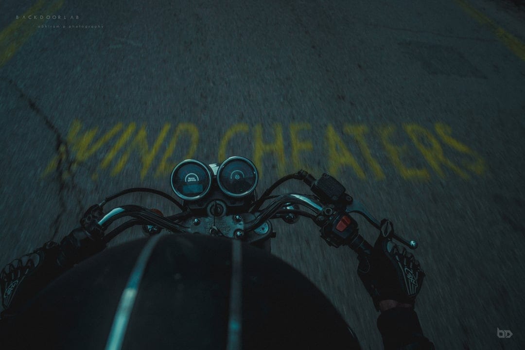 Motorcycle handlebars and dashboard gauges face yellow NO CHEATERS graffiti on a dark asphalt road at night.