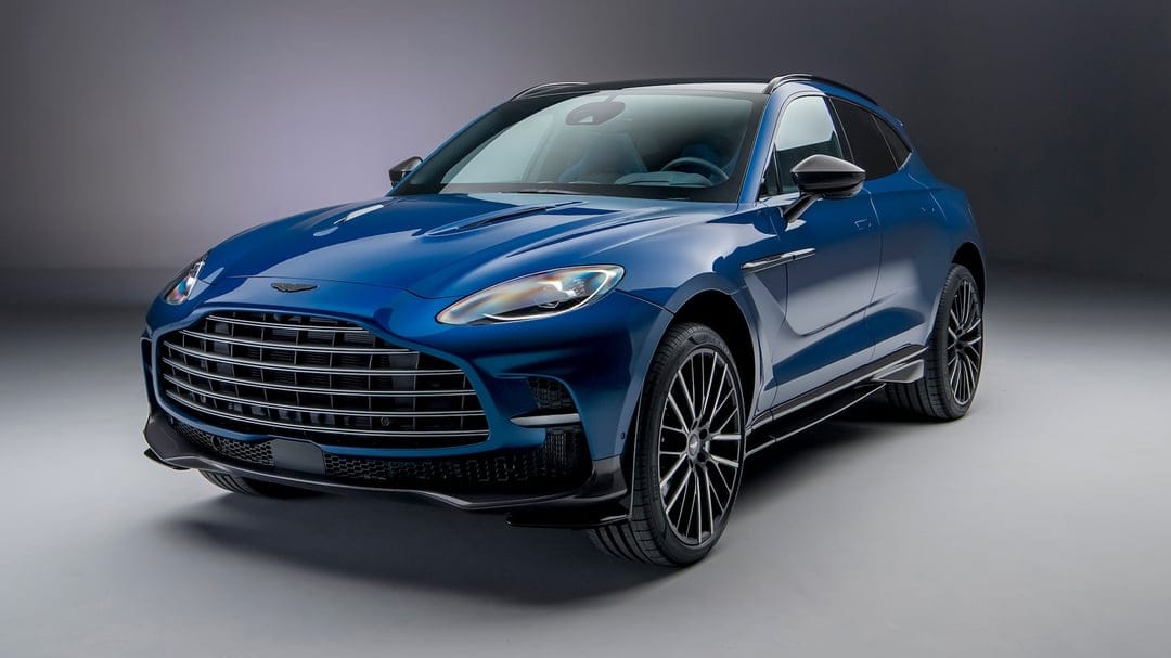 Blue Aston Martin DBX707 SUV with a large black grille and multi-spoke wheels in a grey studio setting.