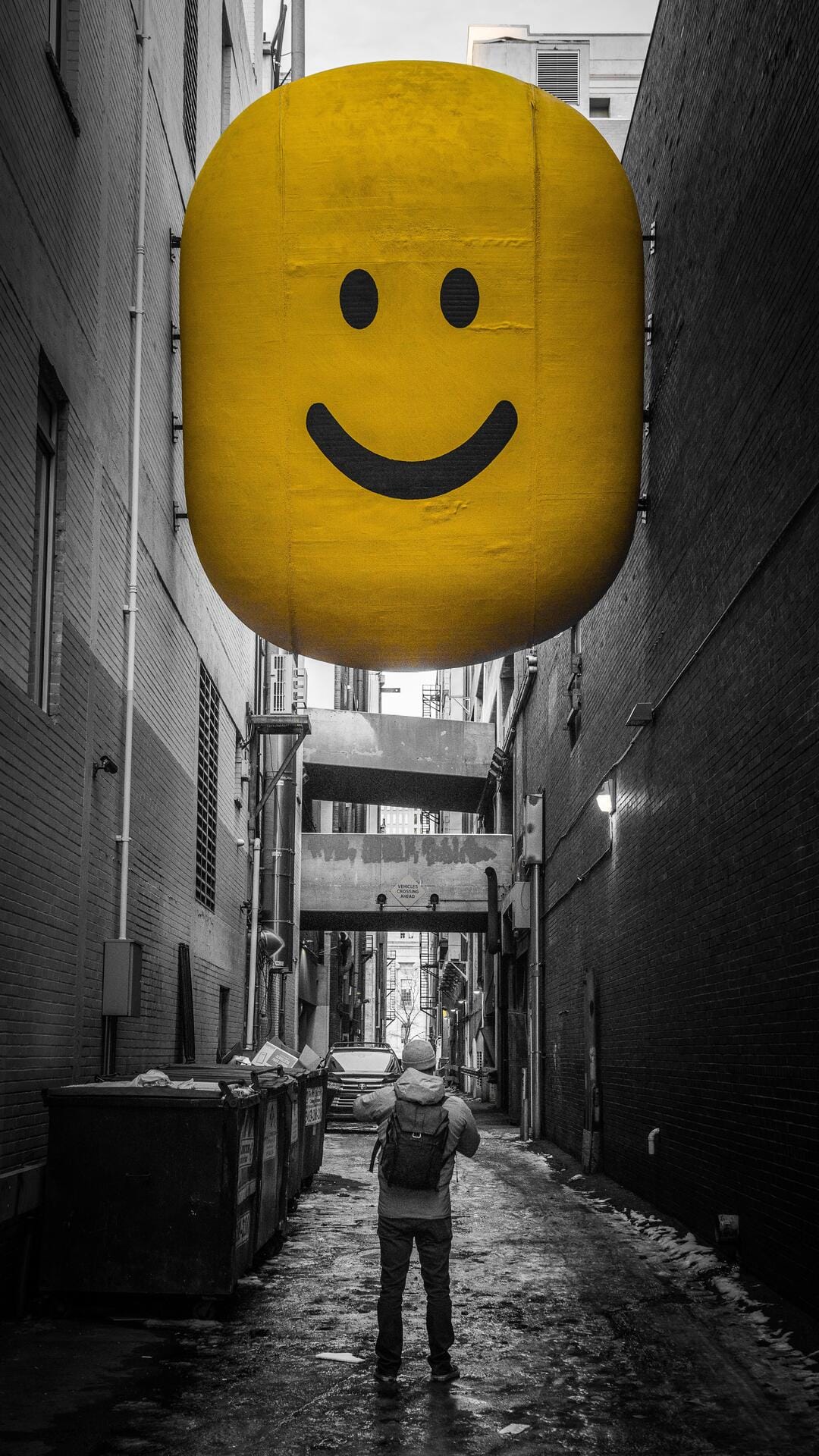 Giant smiling yellow head hovering above a lone person within a narrow black and white urban alleyway.