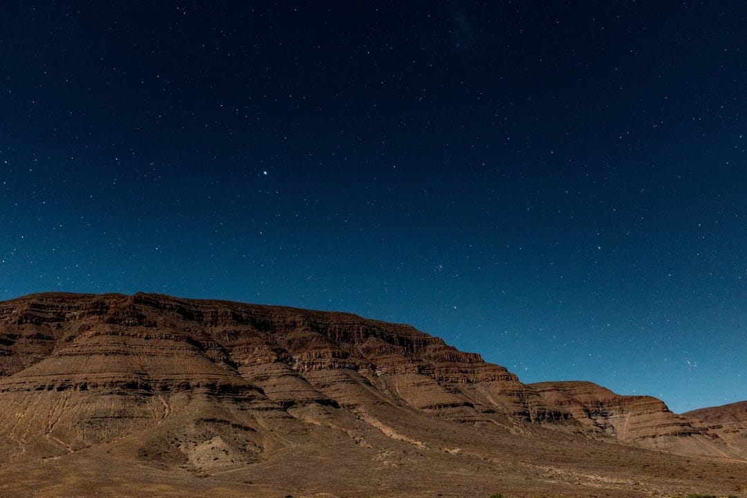 Stratified desert mountains rise toward a clear midnight sky illuminated by countless twinkling stars and nebulae.