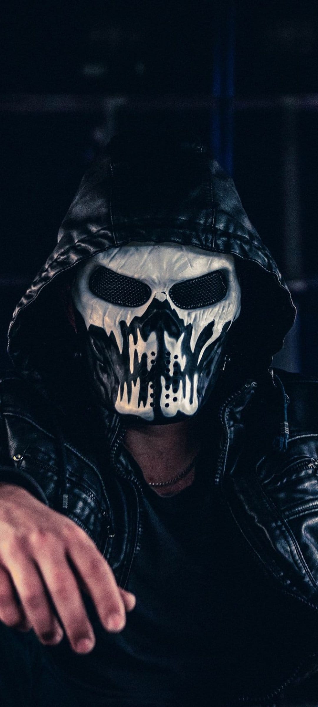 Person in a white skull mask and black leather hooded jacket stares forward against a dark, moody background.