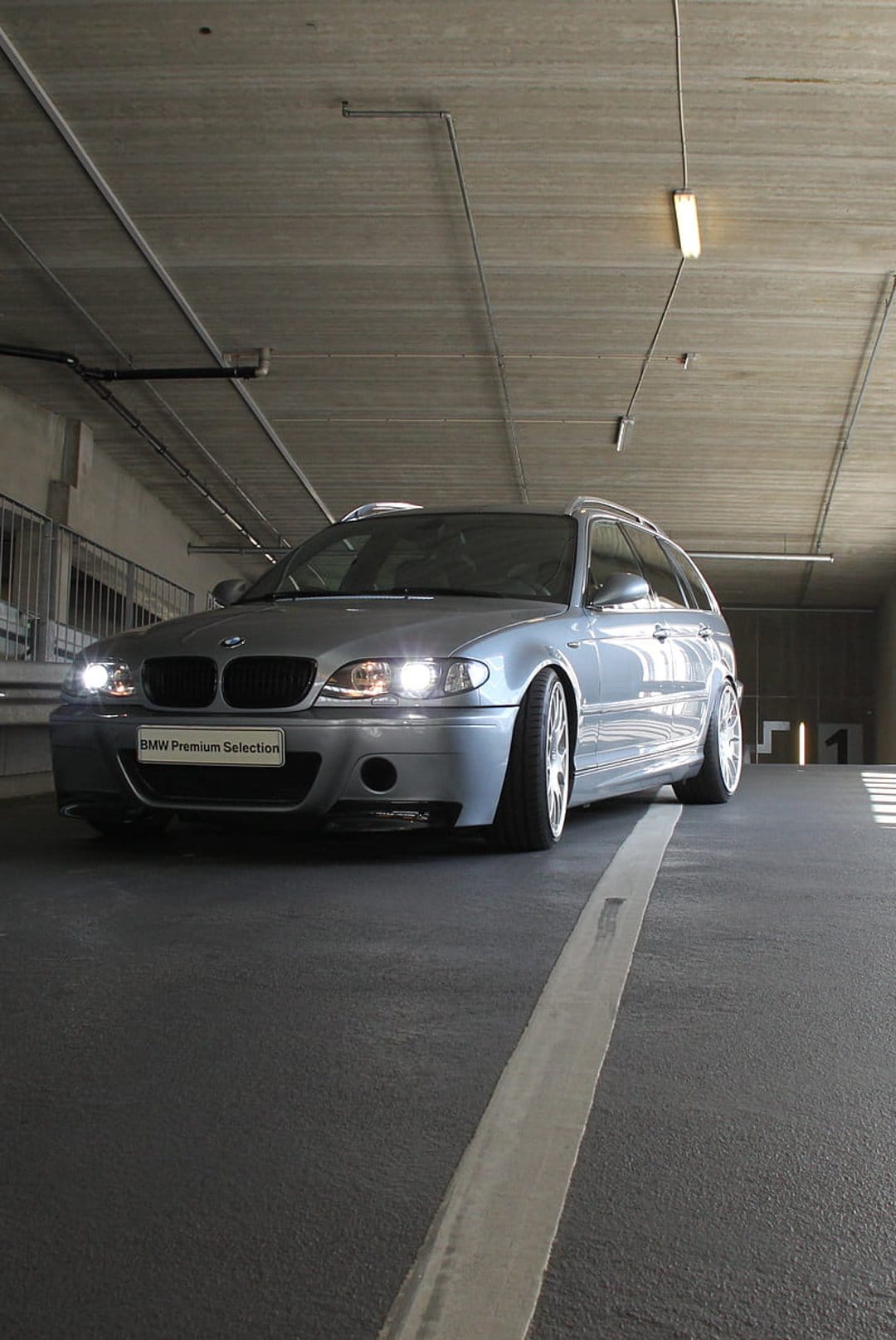 Silver BMW station wagon with glowing headlights parked in a dark concrete garage using a low-angle perspective.