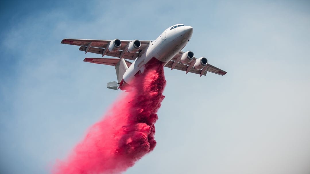White firefighting aircraft releases a massive plume of pink retardant into a clear blue sky during a wildfire.