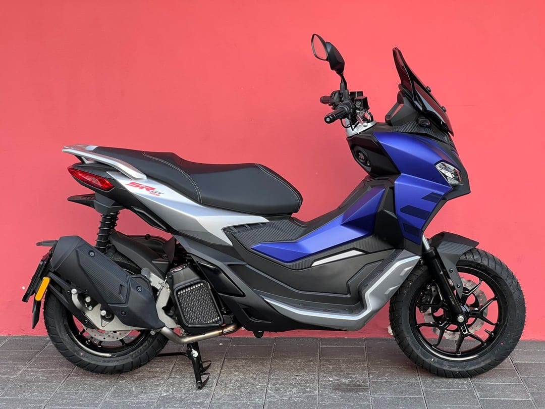 Blue and silver Aprilia SR GT 125 scooter profile against a solid red wall on a grey paved ground.
