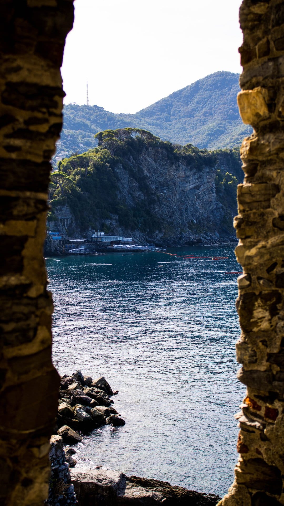 Weathered stone archway framing a view of green Italian hills and the blue Mediterranean sea under bright sunlight.