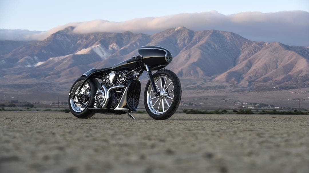 Custom black motorcycle on a desert floor with a backdrop of rugged mountains and soft morning clouds.