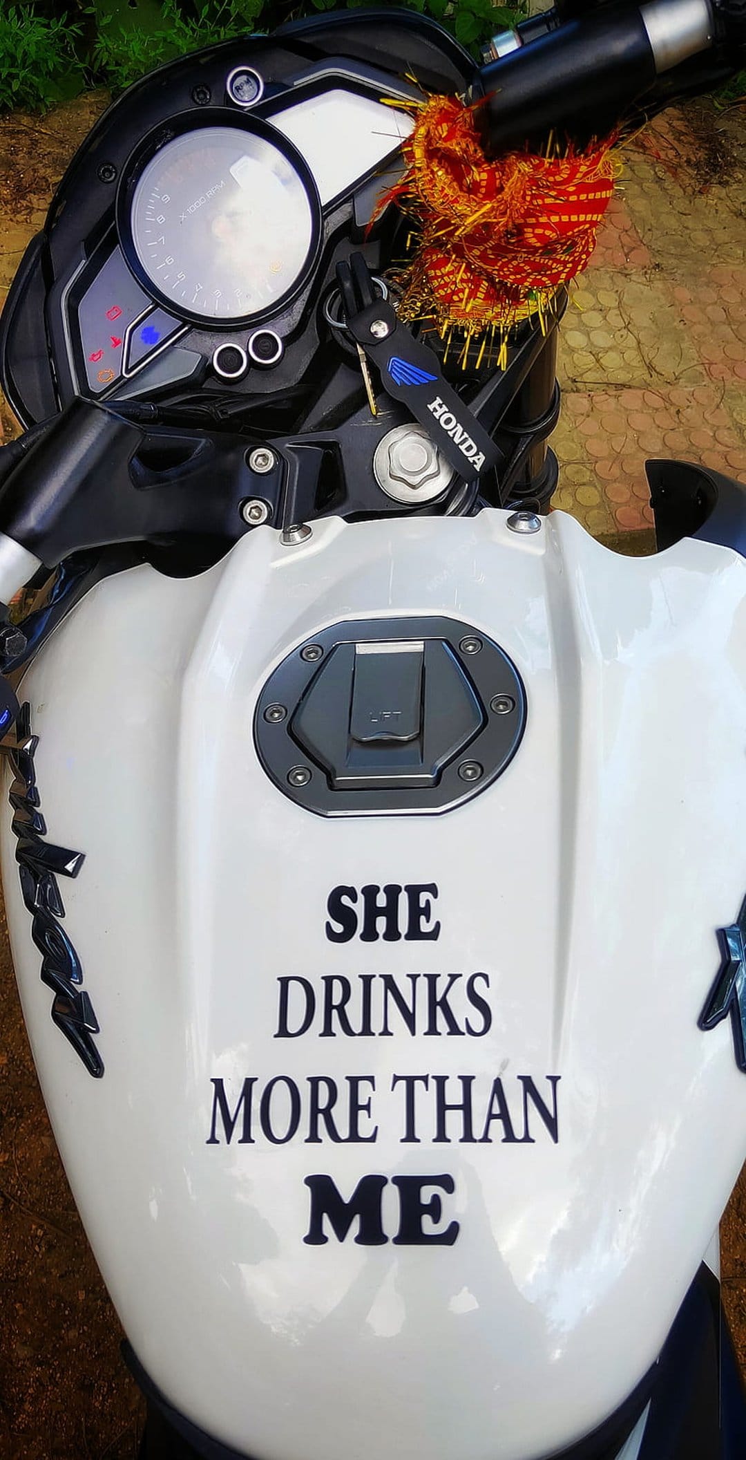 White Honda fuel tank with a black sticker reading She Drinks More Than Me and a red prayer thread on the key.