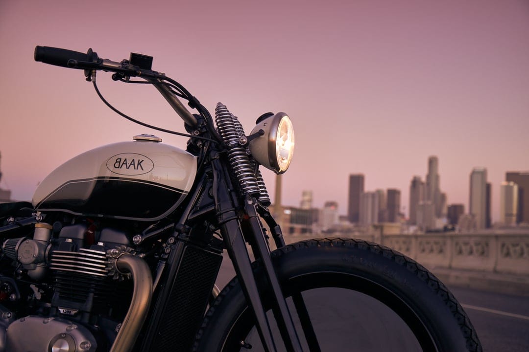 Custom motorcycle with a glowing headlight and cream gas tank parked before a soft pink sunset city skyline.