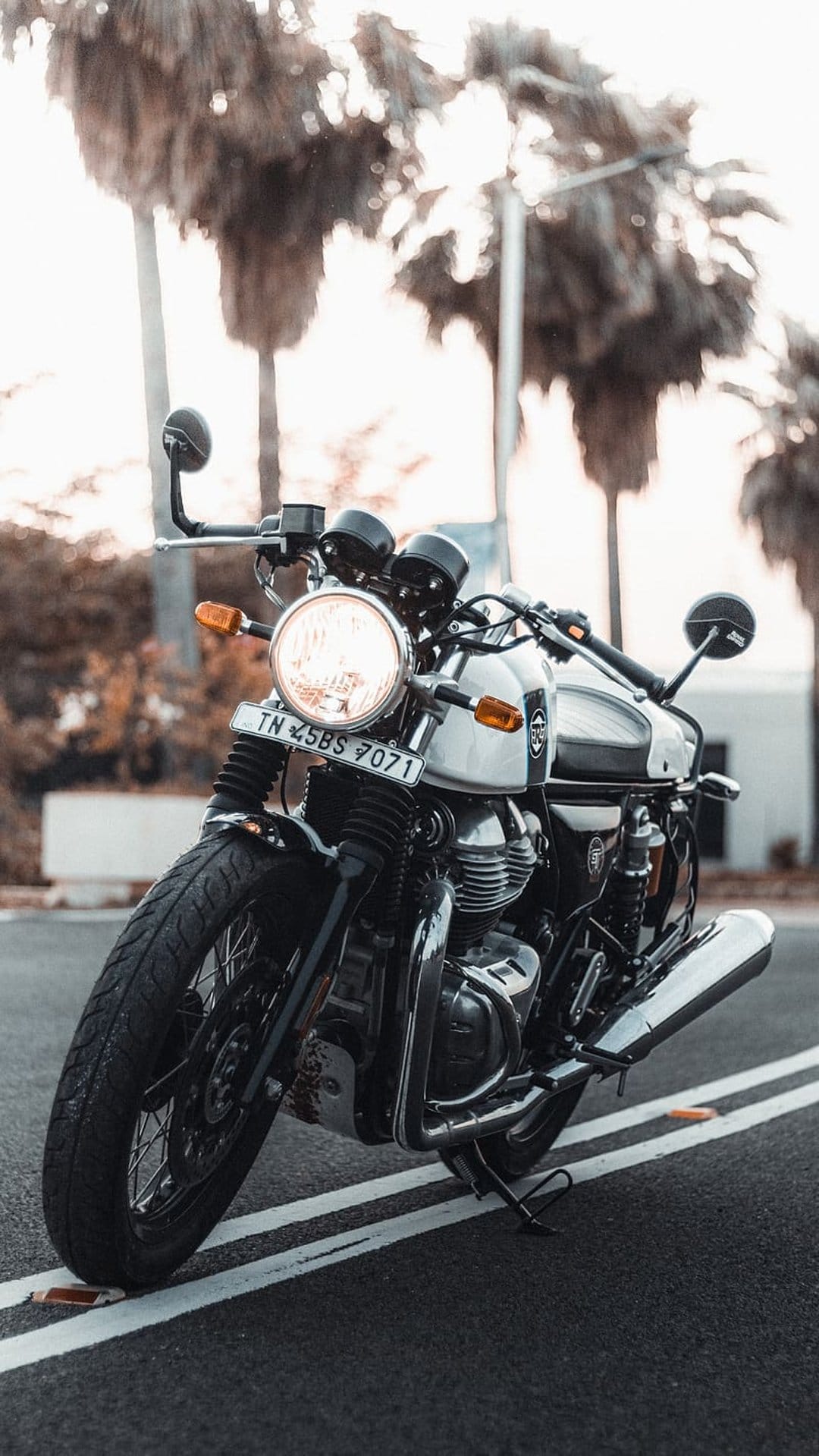 Vintage chrome motorcycle parked on a paved road with tropical palm trees under a bright hazy sky background.