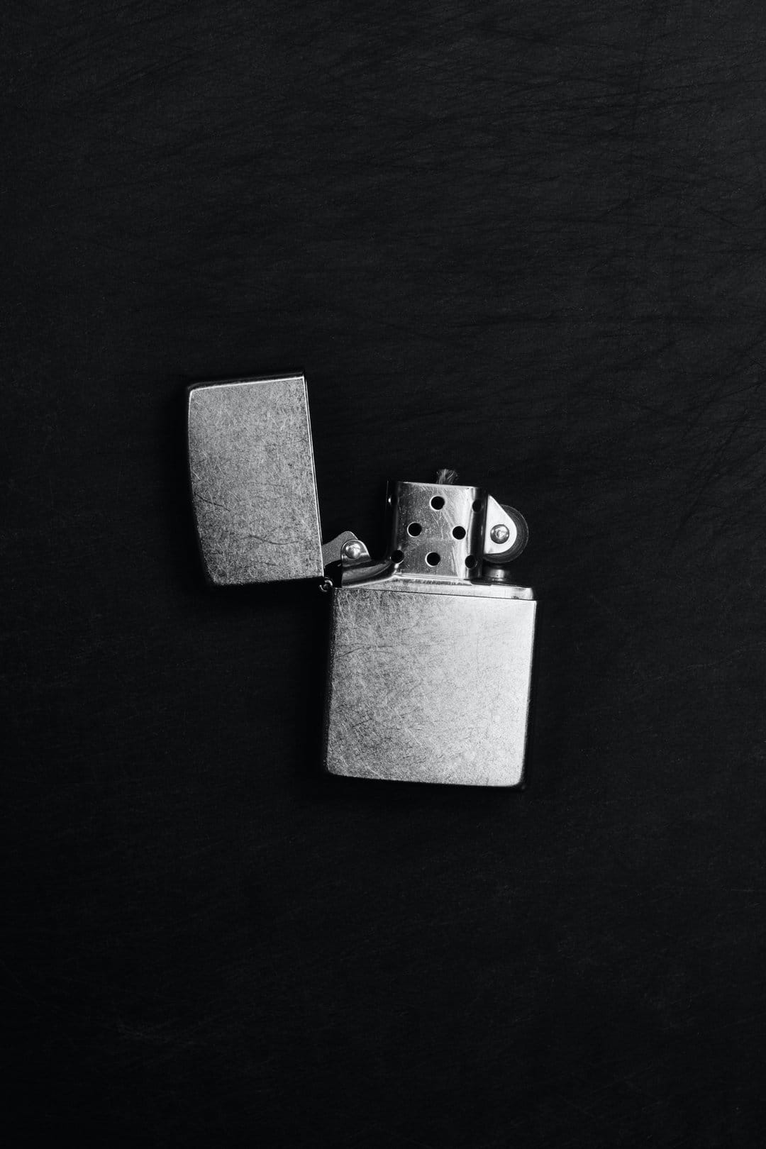 Metallic lighter with an open lid displays its striking wheel on a dark surface in high contrast monochrome.