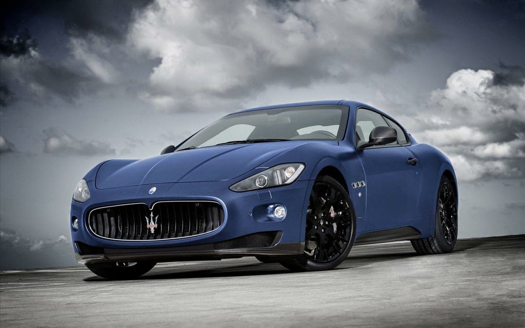 Matte blue Maserati GranTurismo sports car with black rims parked on concrete under a dramatic cloudy sky.