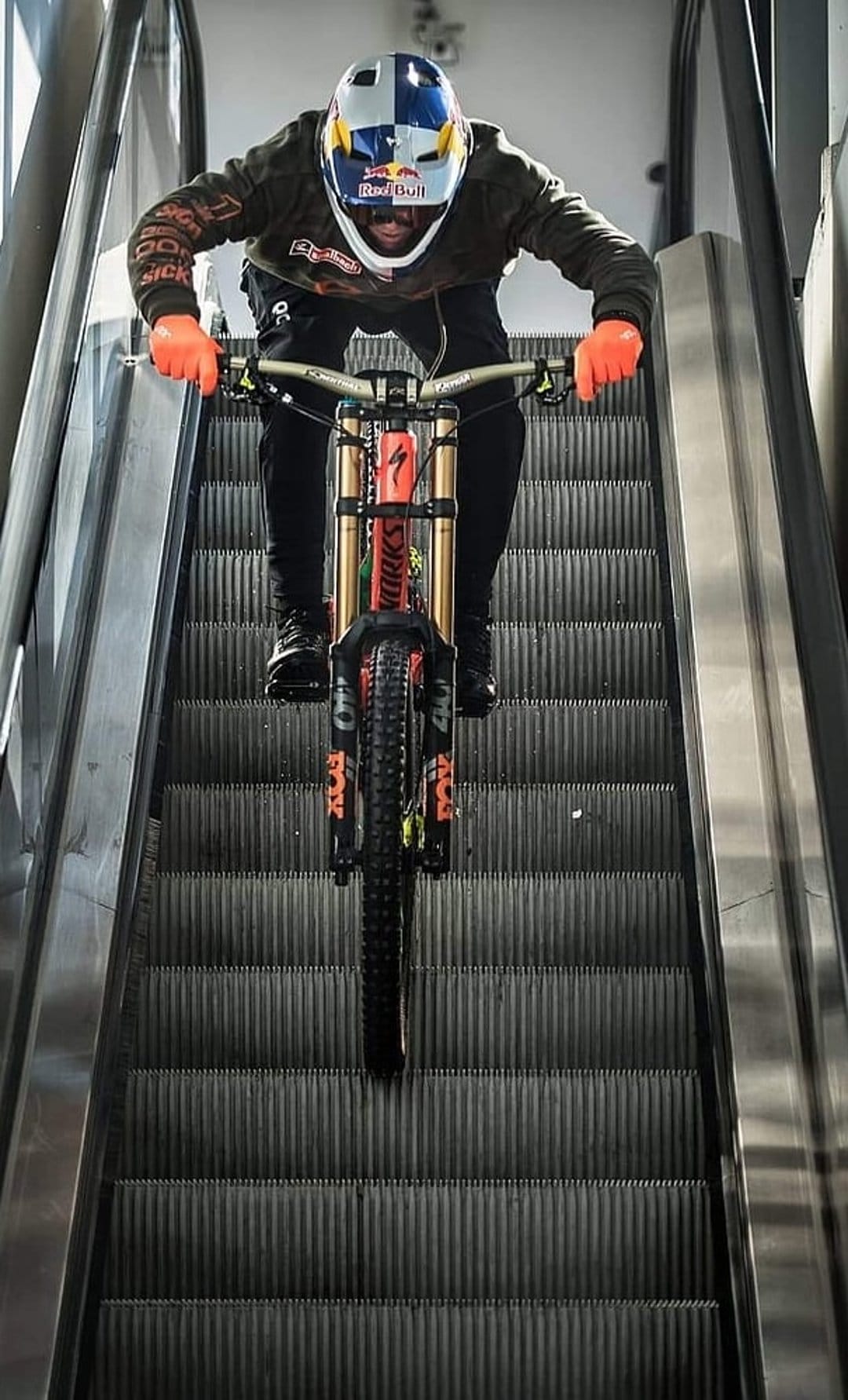 Mountain biker in orange gloves and a helmet rides a full-suspension bike down a metallic escalator.