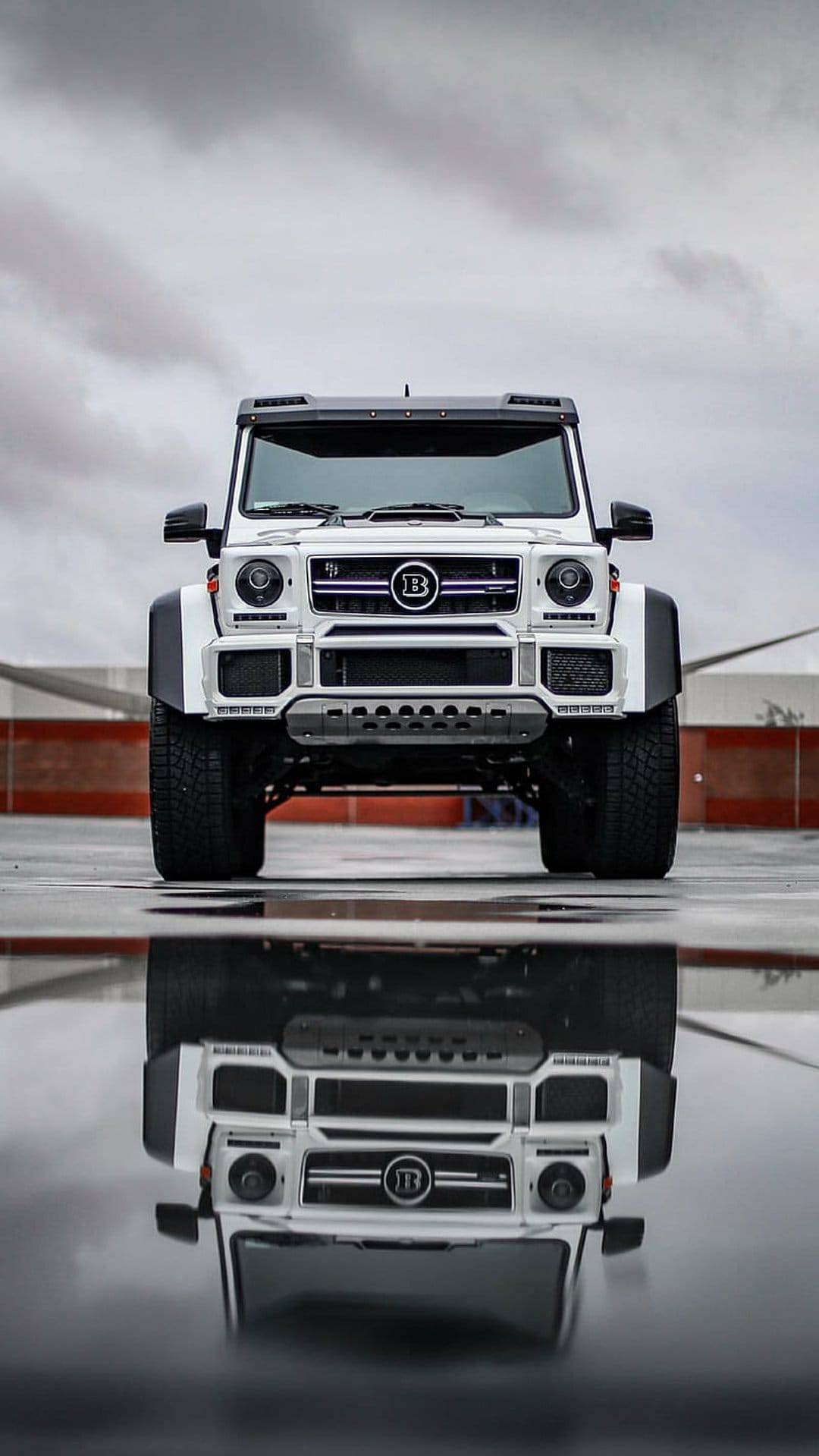 White Brabus G-Wagon SUV reflected in a wet surface under a cloudy sky, captured from a low perspective.