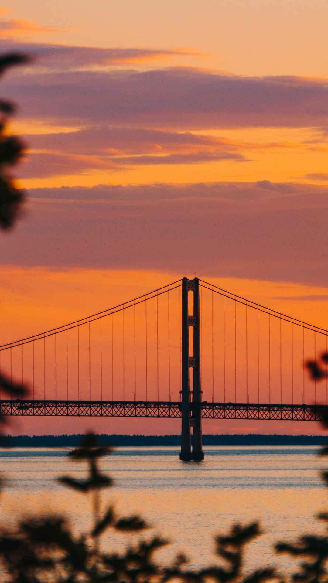Suspension bridge silhouette spanning calm water under a vibrant orange and purple sunset sky with leaf framing.