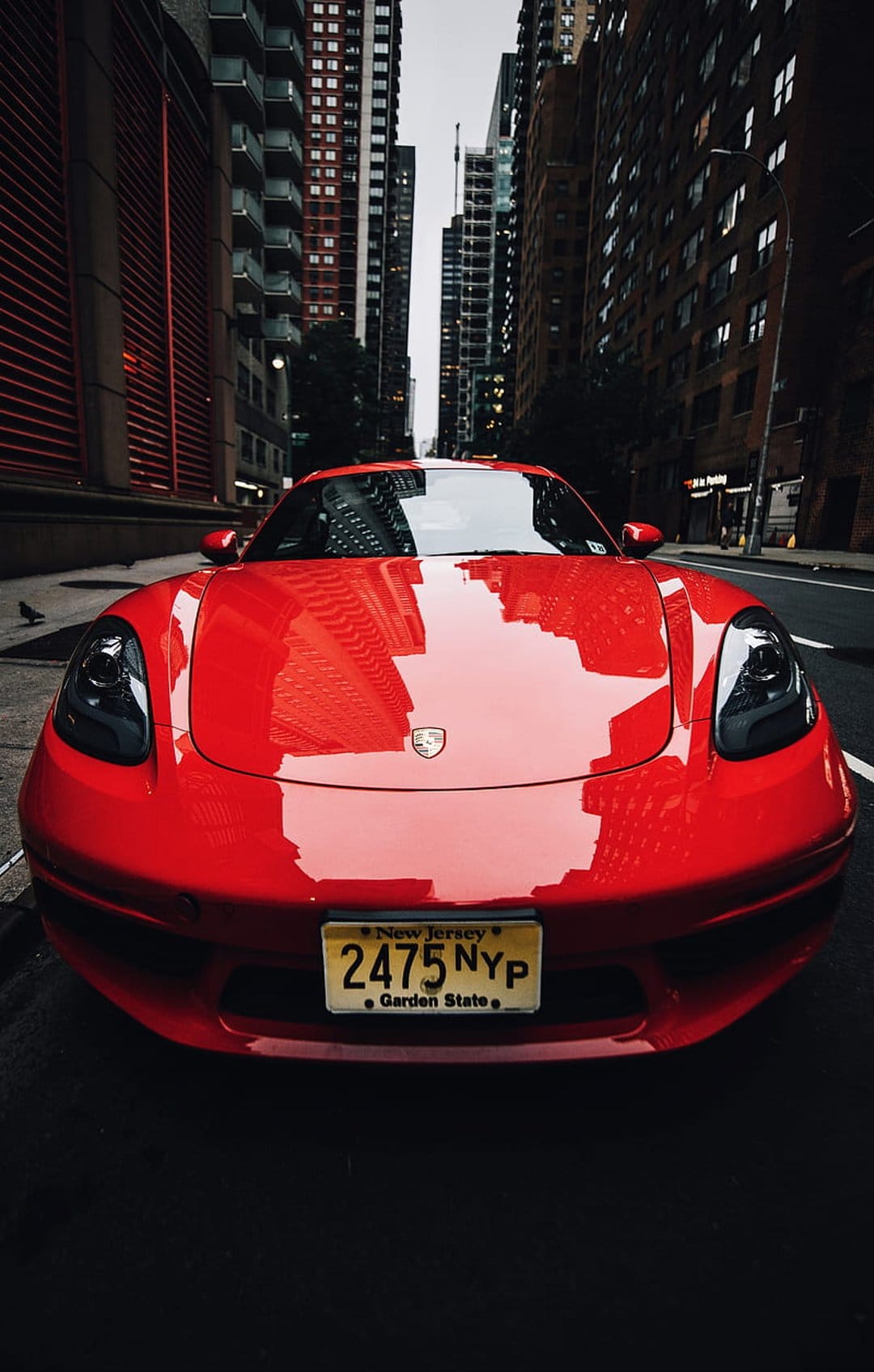 Red Porsche Cayman S parked on a city street with skyscraper reflections on its polished front hood.