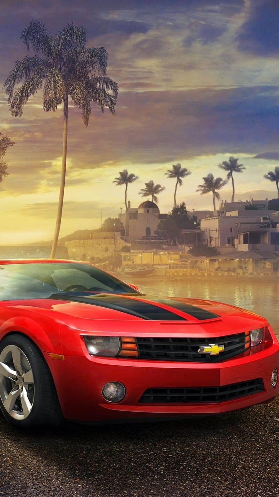 Red Chevrolet Camaro parked on a coastal road with palm trees and a village under a golden sunset sky.