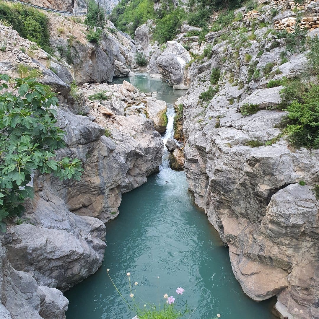Crystalline turquoise river flows between steep grey cliffs and green moss in a vertical canyon landscape.