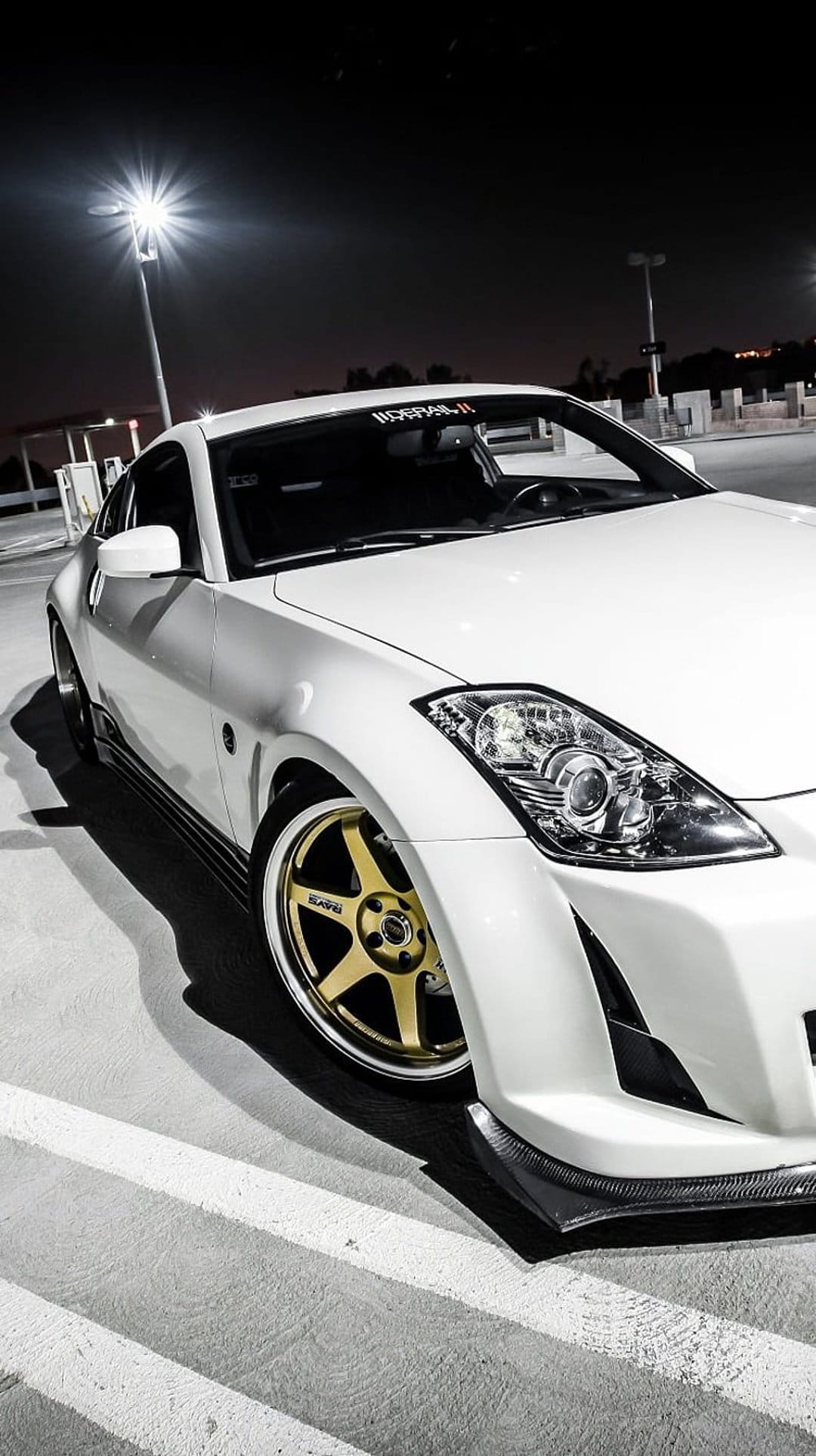 White Nissan 350Z with gold multi-spoke rims parked at night under streetlights in a dark urban parking lot.