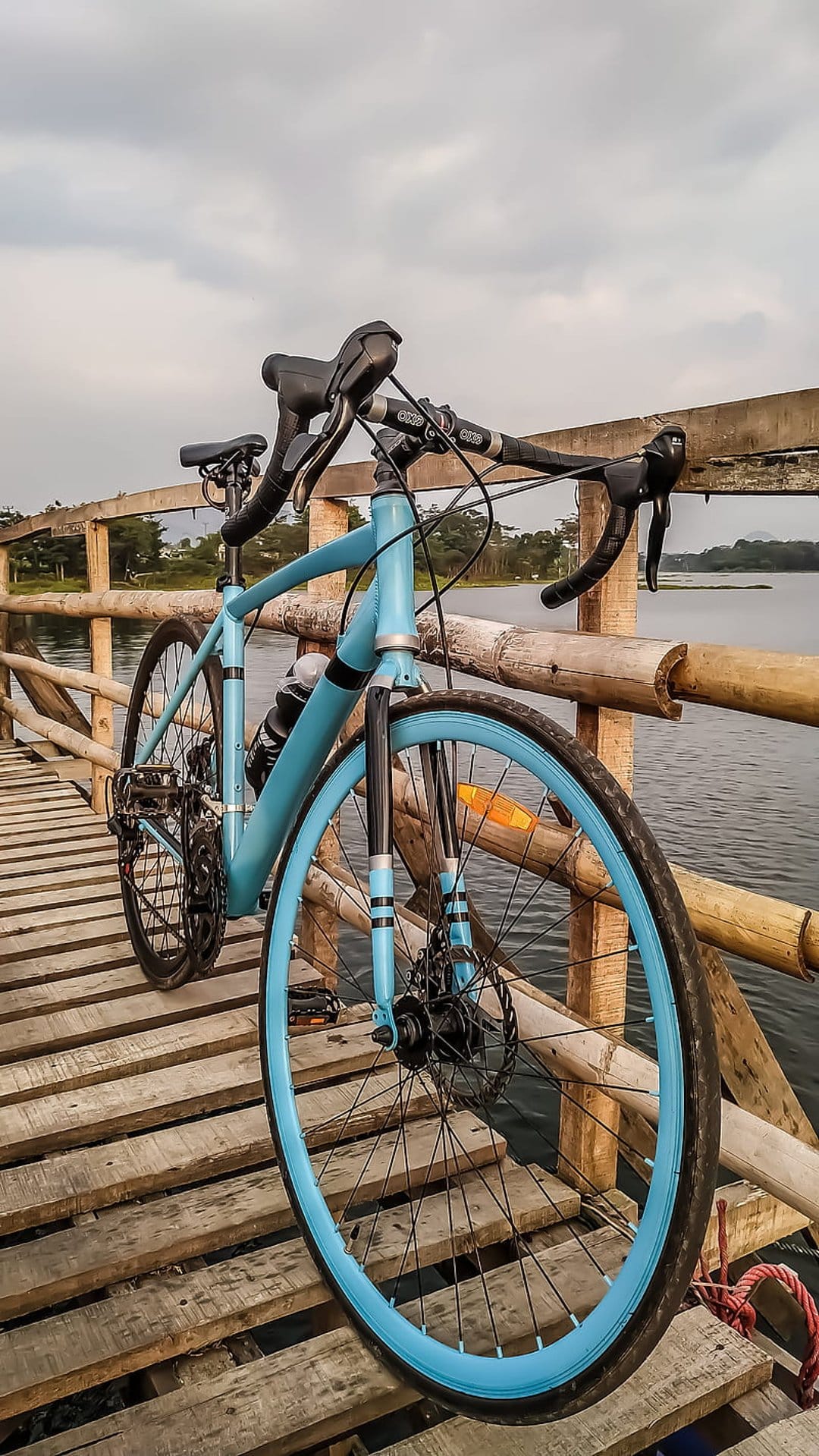 Light blue gravel bike with blue rims parked on a weathered wooden boardwalk beside a calm lake under clouds.