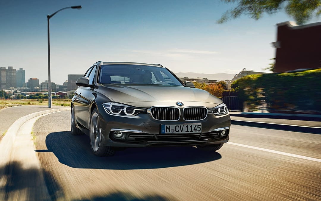 Grey BMW 3 Series Touring driving on a sunlit urban road with a city skyline and clear blue sky in the background.