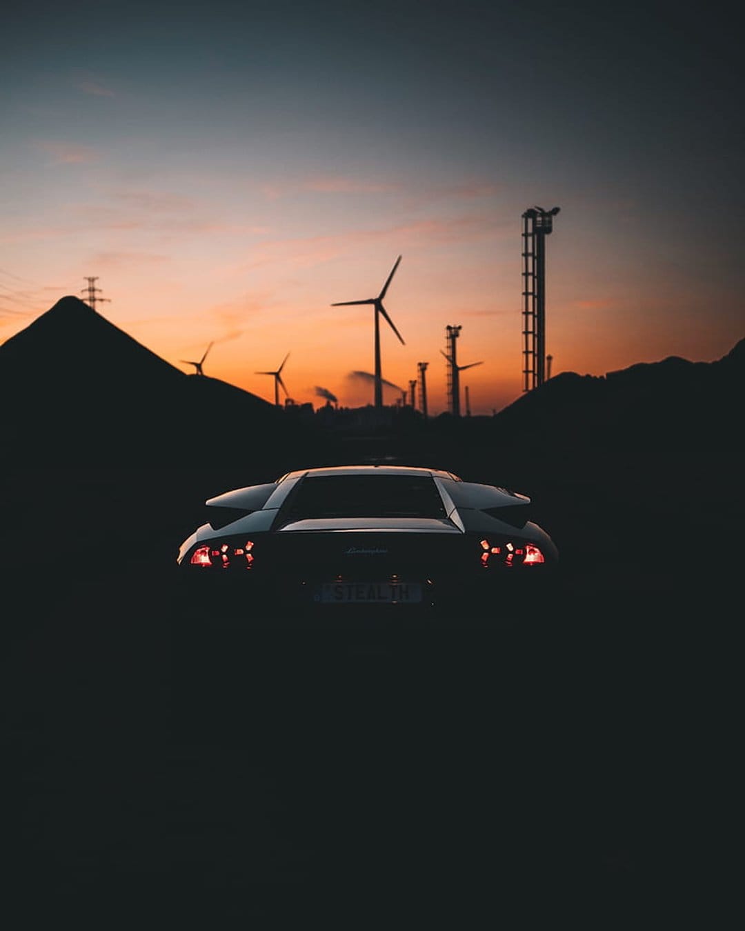 Lamborghini silhouette with glowing red taillights parked near wind turbines under a dramatic sunset sky.
