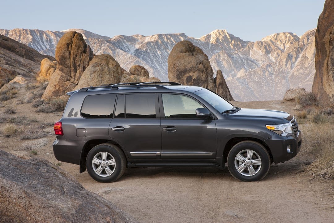 Dark grey SUV on a dirt path surrounded by towering rocks and snow-dusted mountains under a clear sky.