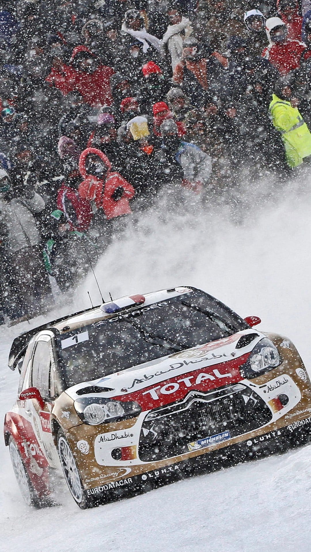 Citroen DS3 WRC rally car drifting through deep snow on a winter track with spectators and falling snowflakes.