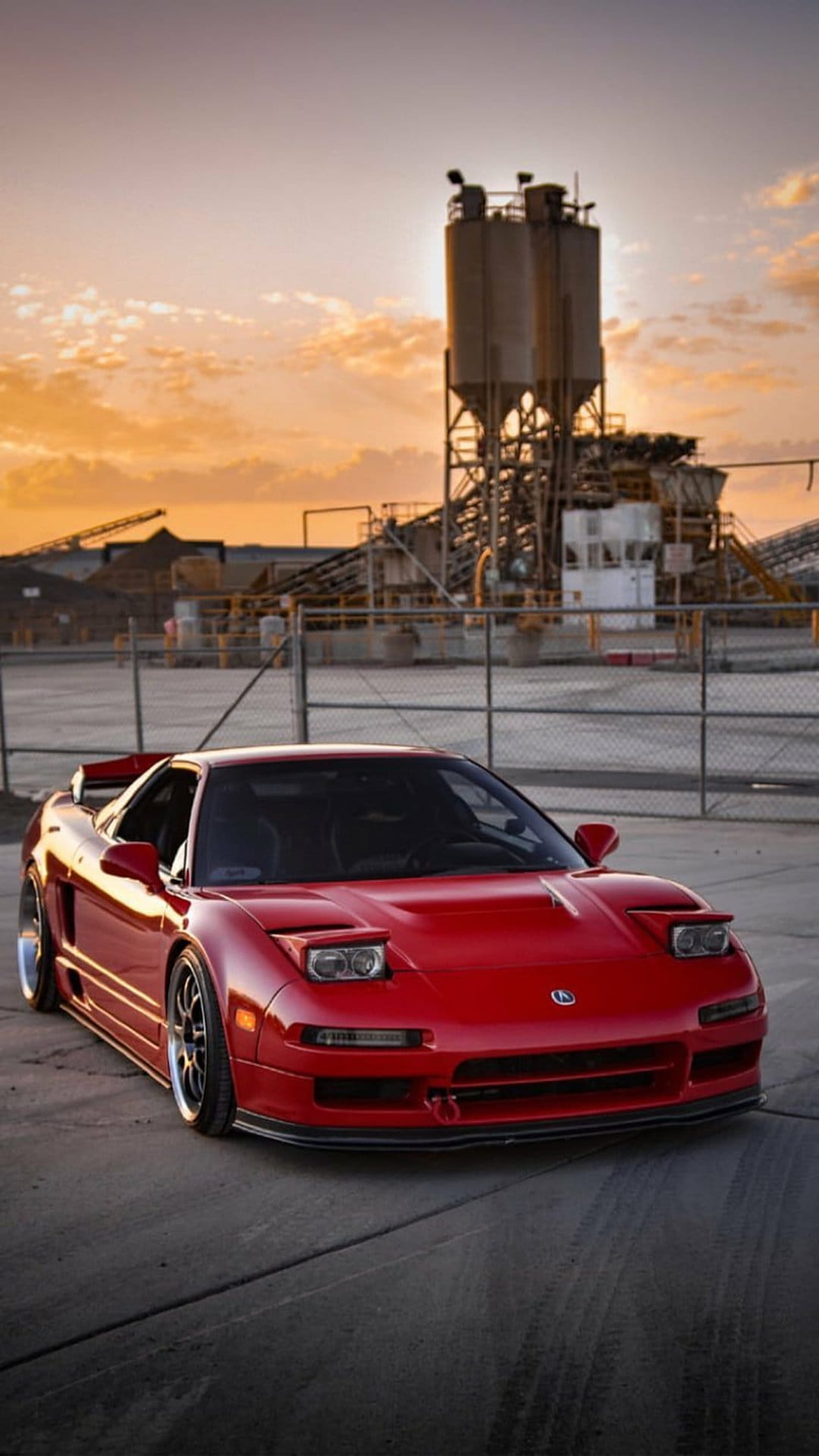 Red Acura NSX with pop-up headlights parked at a sunset industrial site with silos in a low-angle vertical shot.