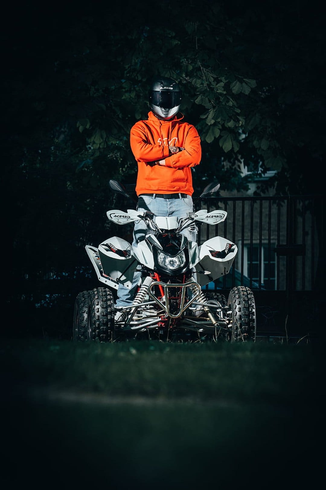 Person in an orange hoodie and helmet stands on a white ATV against a dark night background with a wooden fence.