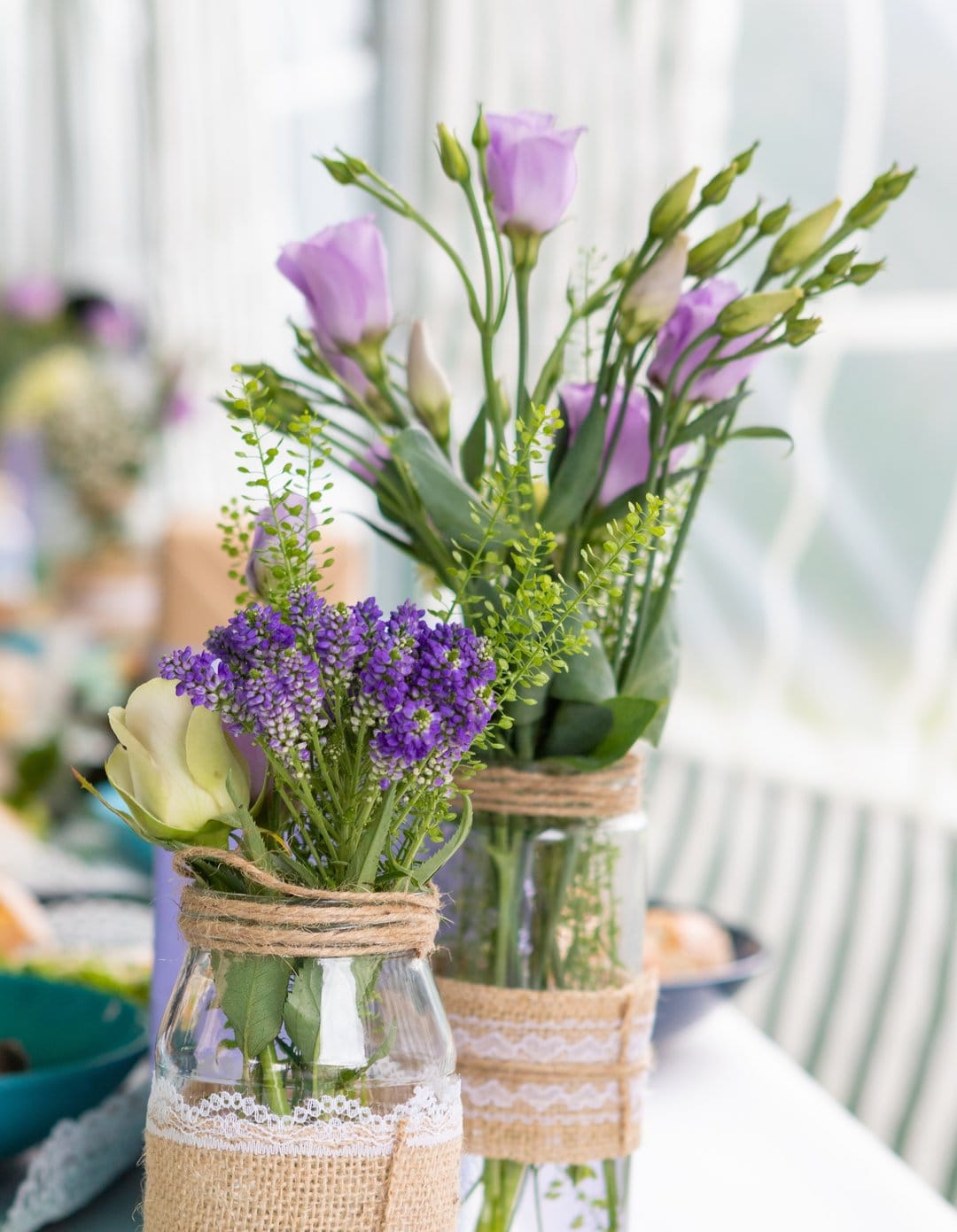 Purple flowers and a white rose in glass jars wrapped in burlap and lace set against a soft-focus background.