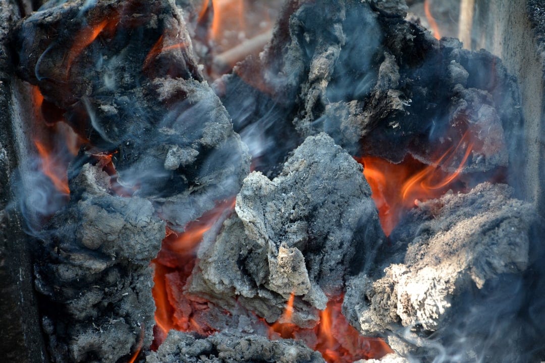 Glowing orange embers and charred black wood emit thin trails of grey smoke in a detailed macro vertical scene.