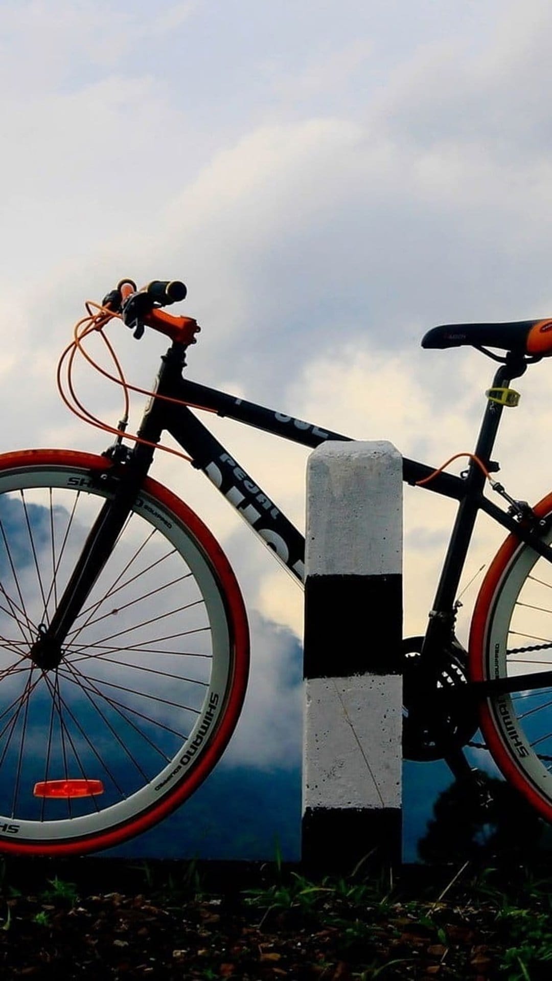 Black road bike with orange tires leaning on a striped pole against a backdrop of mountains and a cloudy sky.